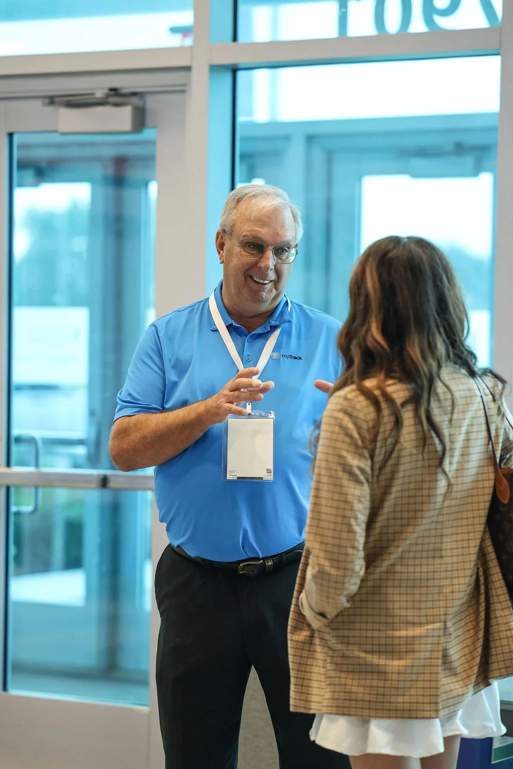 A man in a blue shirt with a name badge is talking to a woman in a beige blazer at an indoor event, near glass doors.