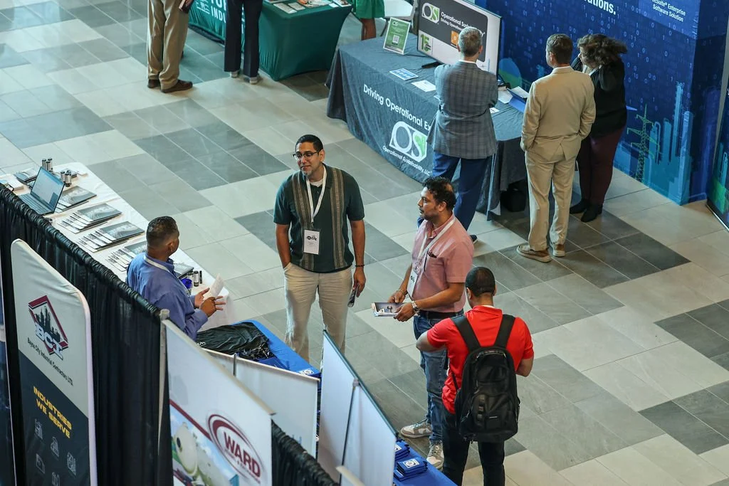 People attending a trade show or conference, engaging in conversations and exploring booths with banners and informational displays in an indoor venue.