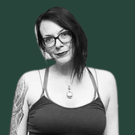 Black and white photo of a woman with glasses, tattooed arm, wearing a crop top and skirt, standing against a dark green background.