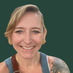 A smiling woman with short blonde hair, a tattoo on her left arm, wearing a light blue overall dress over a white top, standing against a solid green background.