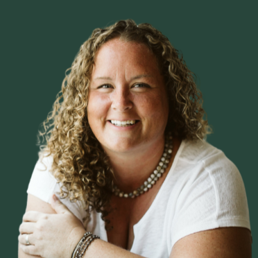 A woman with curly blonde hair smiling, wearing a white shirt and a pearl necklace, against a green background.
