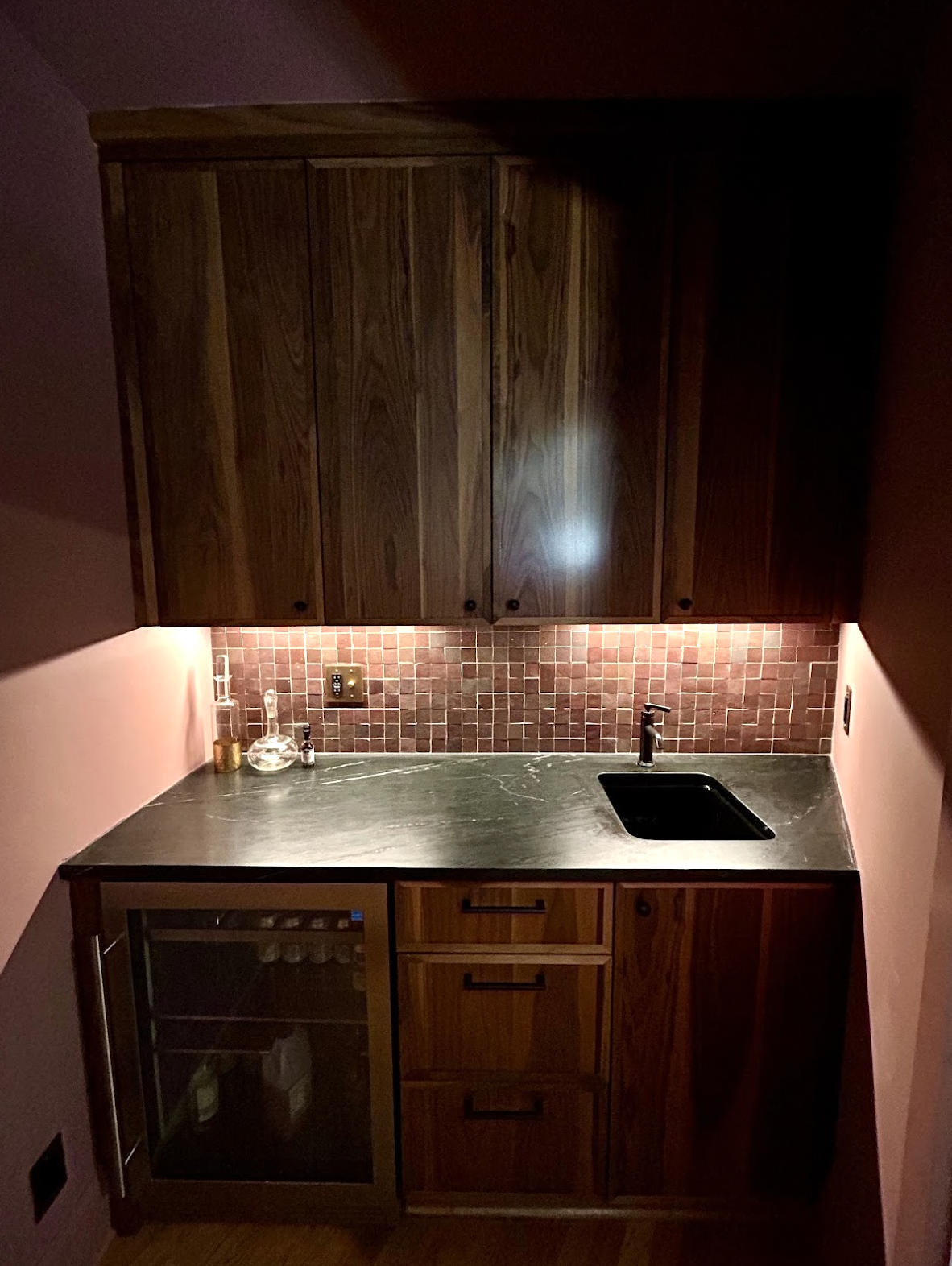Custom bar backsplash featuring warm Zellige tile, under-cabinet lighting, dark countertops, wood cabinetry, bar sink, and precision tile installation by Parker’s Precision Tile.