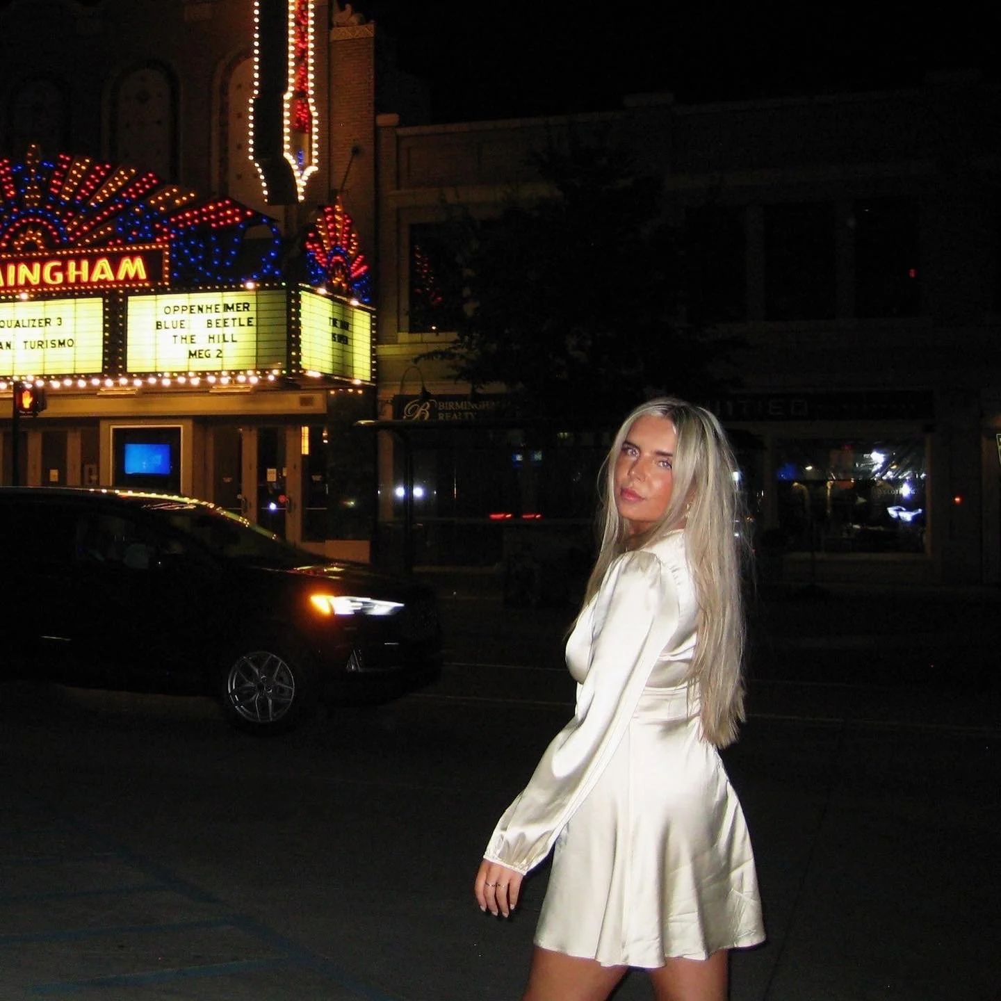 A woman with long blonde hair in a white satin dress standing on a dark street at night with bright carnival lights and a marquee in the background.