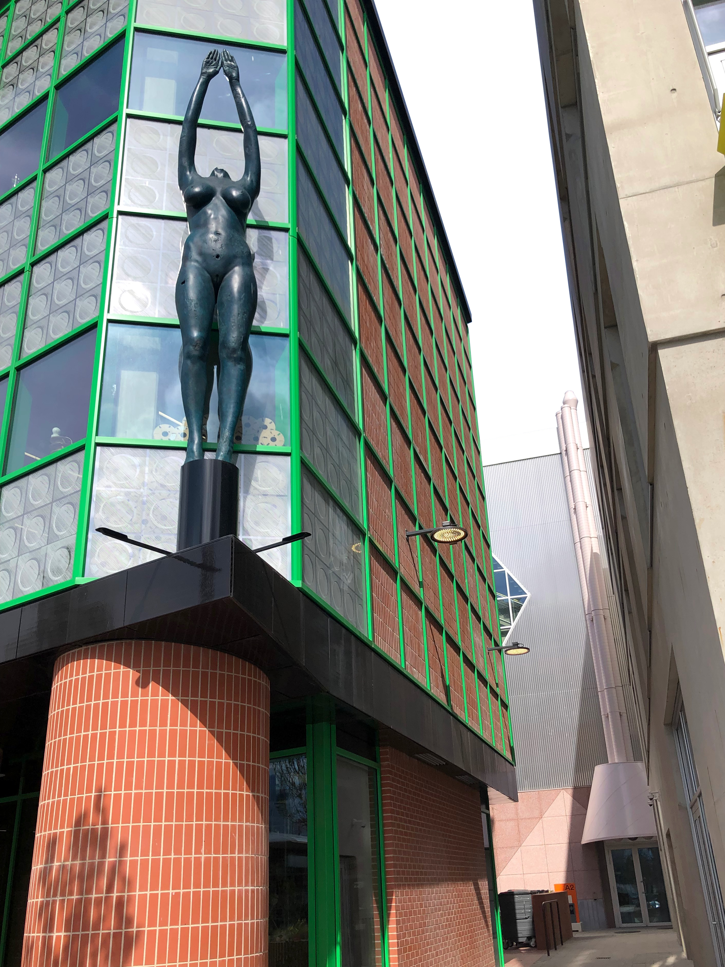 A tall black statue of a nude woman with arms raised above her head by Damien Hirst is mounted outside on the corner of a modern building with green window frames and brick accents.