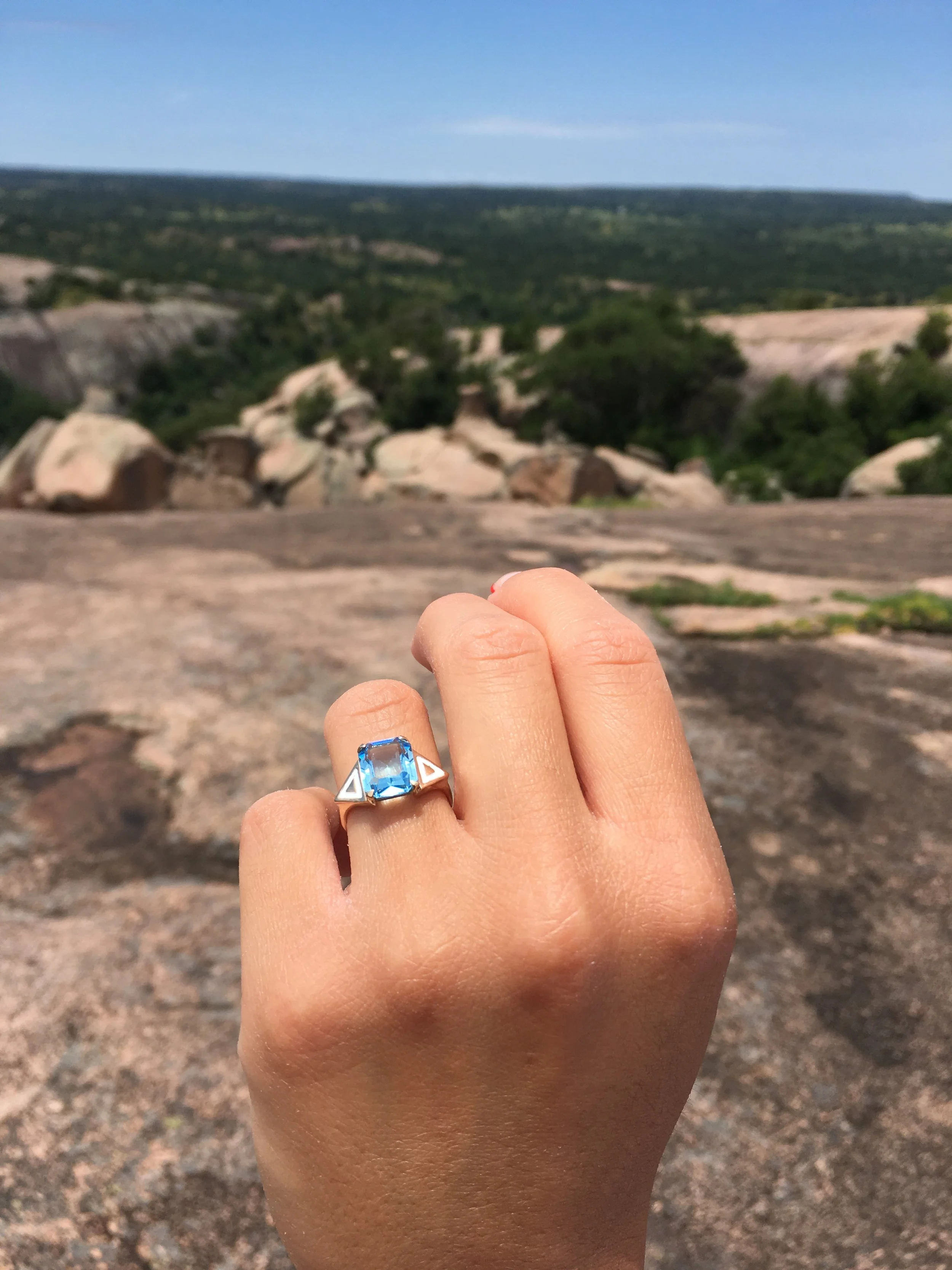 Hand with a silver ring featuring a large blue gemstone, held against a landscape of rocks, trees, and distant horizon under a blue sky.