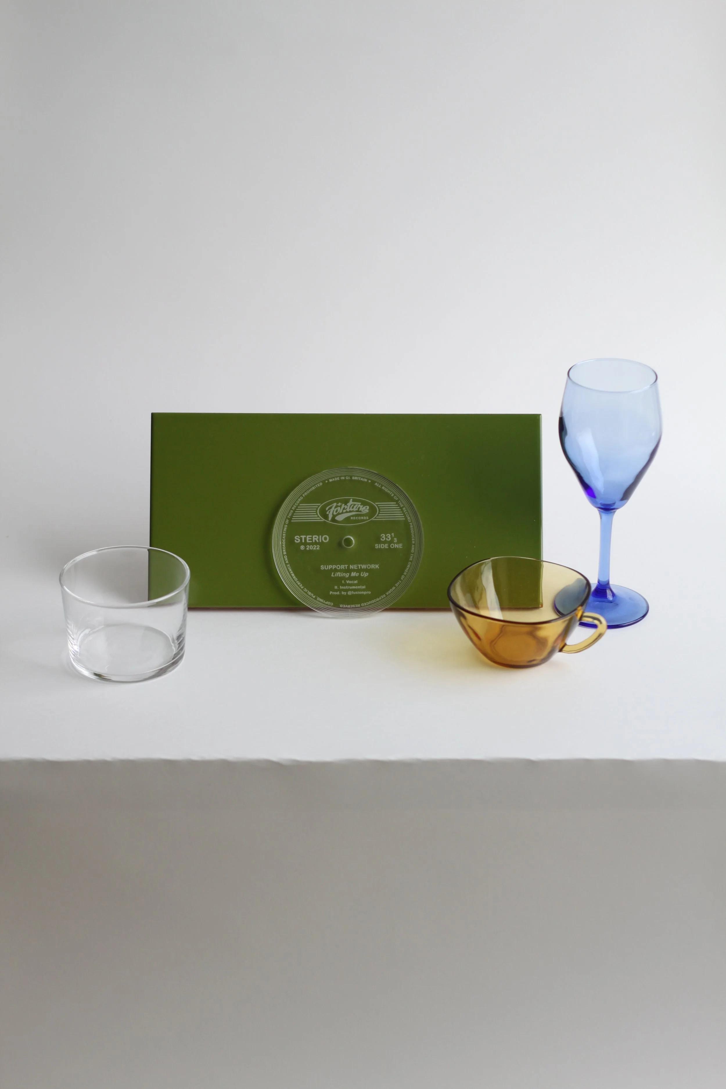 A collection of glassware and a green record sleeve with a record inside. The glassware includes a clear short tumbler, a BOJ blue wine glass, and a brown whisky glass with a gold rim.