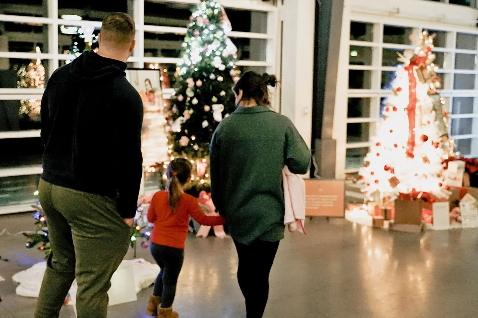 A family of three, a man, woman, and young girl, looking at decorated Christmas trees in a festive indoor setting.
