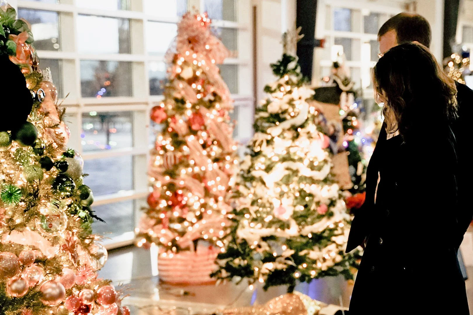 People standing near decorated Christmas trees with lights in a festive indoor setting.