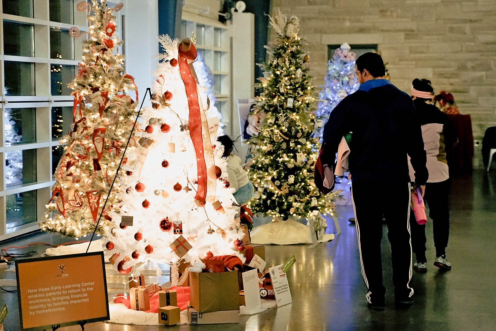 People walking past decorated Christmas trees with lights, ornaments, and presents in a festive indoor setting.