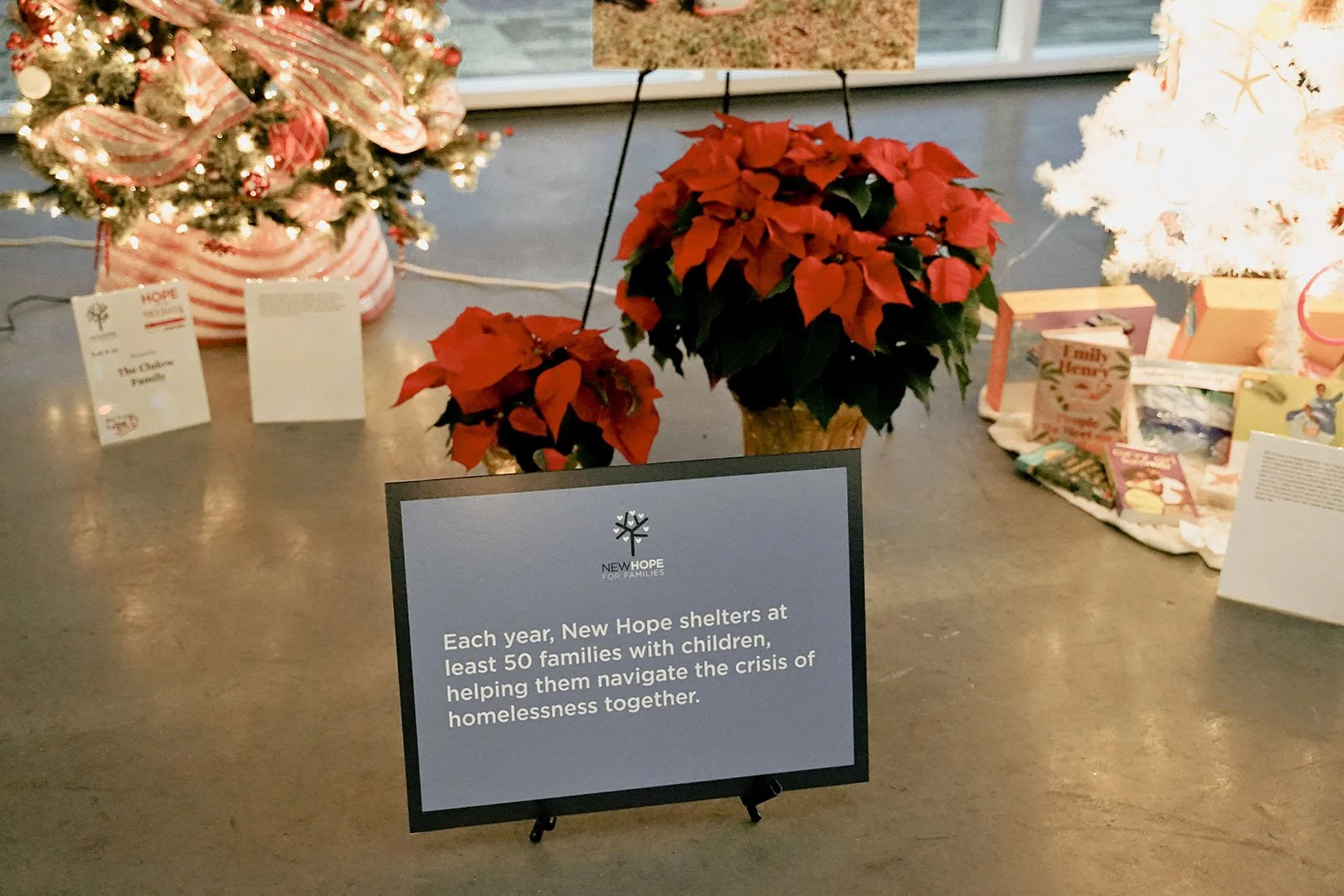 Display table with two poinsettia plants in pots, Christmas trees decorated with ornaments and ribbons, and holiday greeting cards along with a sign that explains the work of New Hope shelters in helping families with children facing homelessness.
