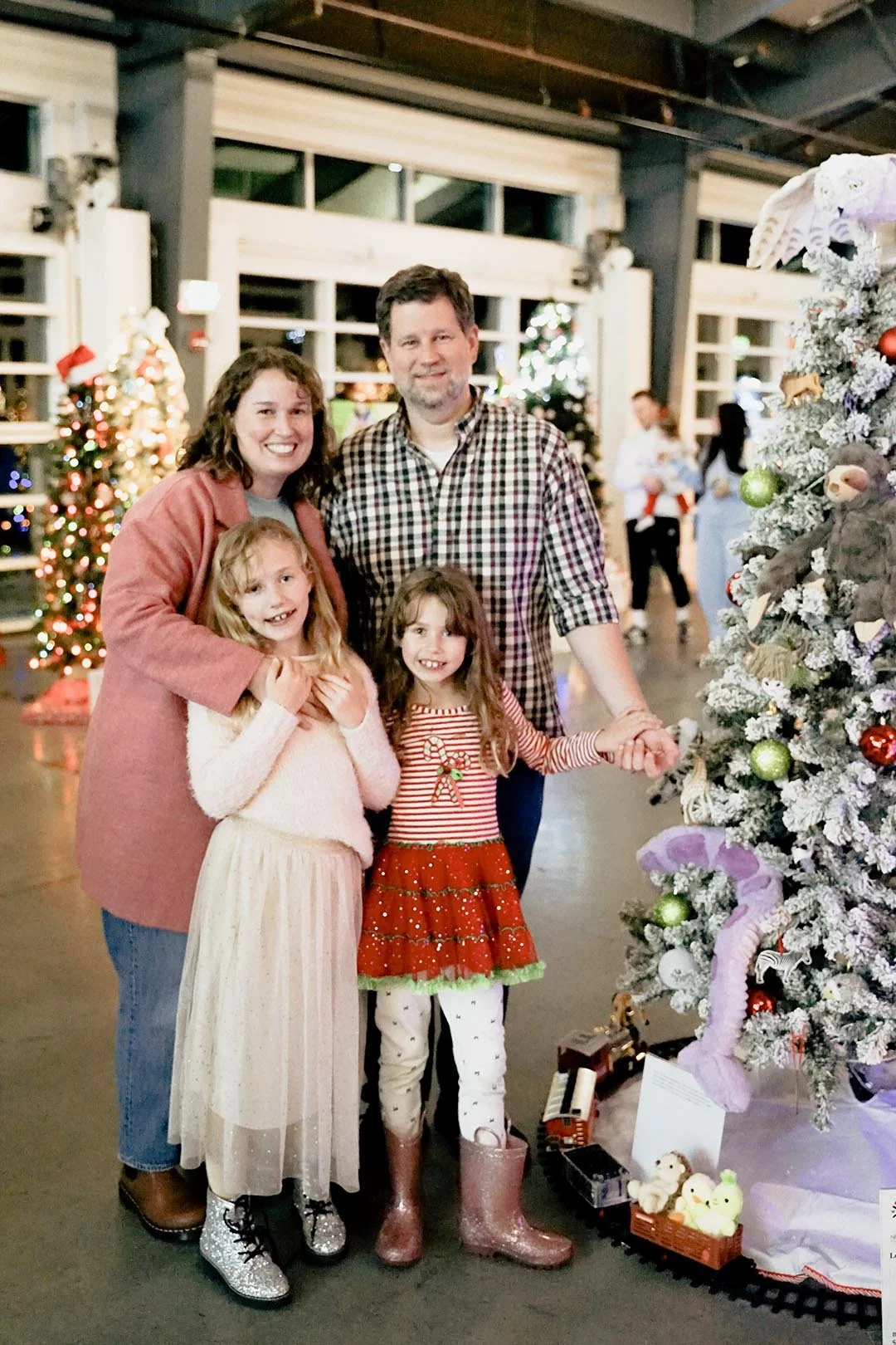 A family of four standing in front of a decorated Christmas tree at an indoor holiday event. The mother is wearing a pink coat, the father a plaid shirt, one daughter in a cream-colored dress, and the other in a red and green holiday-themed dress wit