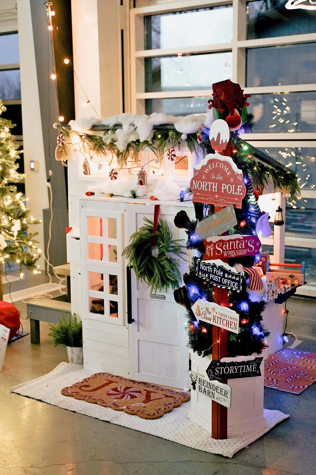 Christmas display with a white wooden house decorated with Christmas lights, garland, and cotton snow, and a signpost with holiday-themed signs like 'Welcome to the North Pole,' in a room with large windows and Christmas trees.