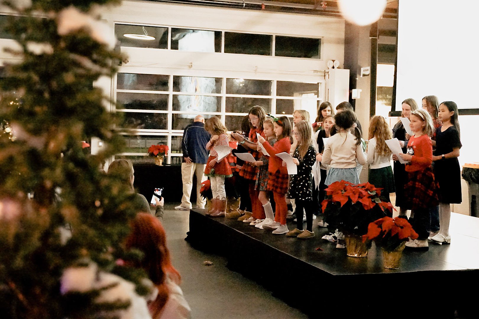 Children singing on stage during a Christmas event, decorated with poinsettias, with an audience watching and a blurred Christmas tree in the foreground.