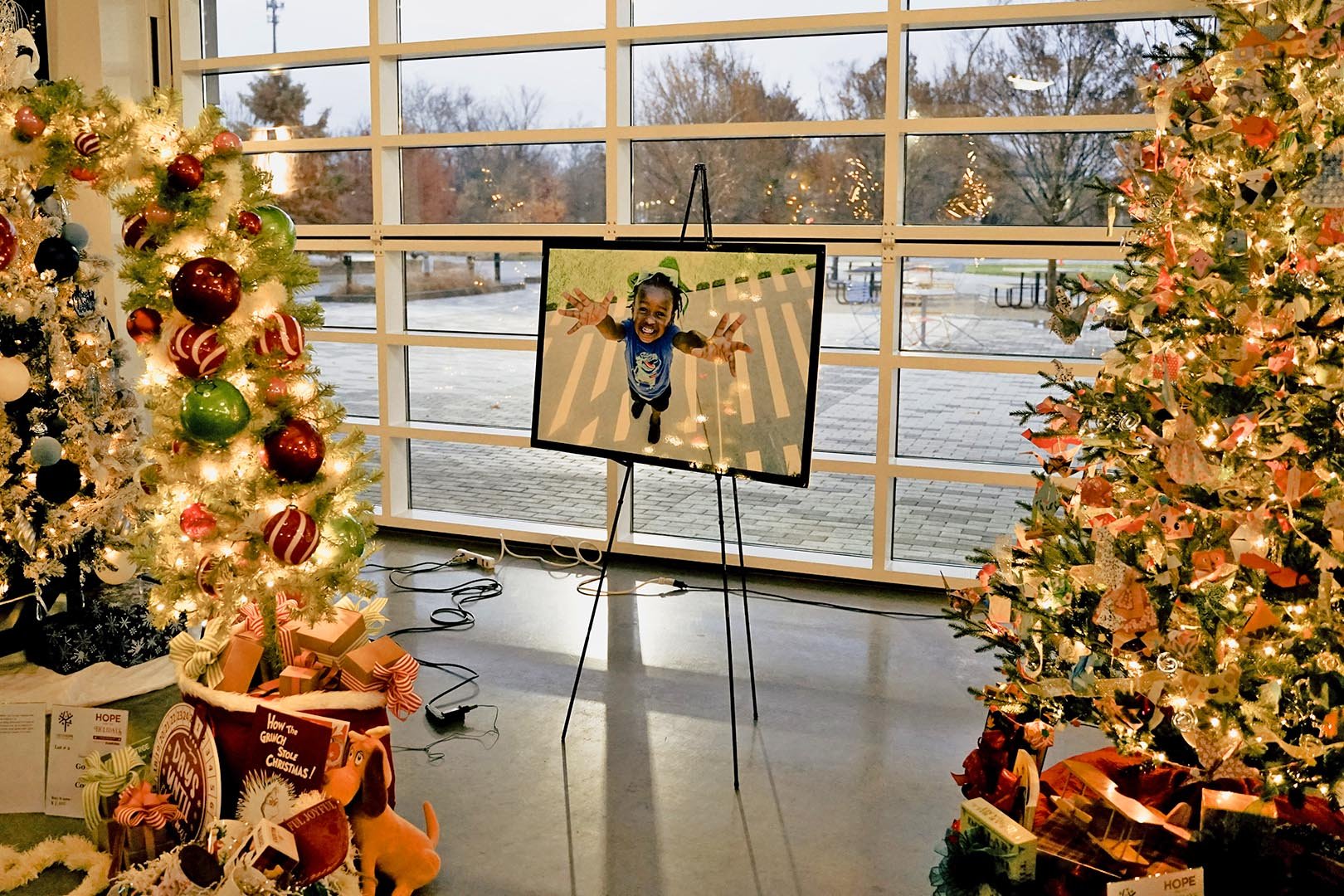 A decorated Christmas display with two Christmas trees with lights and ornaments. In the center, a screen on a stand shows a young girl reaching towards the camera with her arms outstretched. There are wrapped presents, a plush toy, and holiday-theme