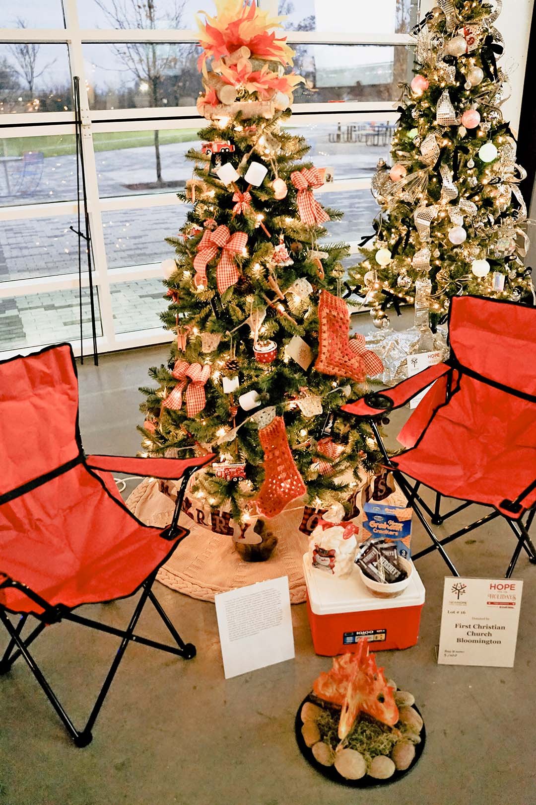 Two decorated Christmas trees surrounded by red chairs, with a window showing an outdoor scene and Christmas decorations including plaques, a snowman, and a fox sculpture on the floor.