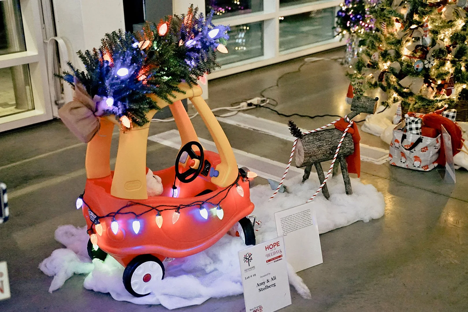 A children's Christmas display featuring a red toy vehicle with a Christmas tree decorated with lights and ornaments, placed on cotton resembling snow, with a wooden reindeer figure nearby and a Christmas tree in the background decorated with lights 