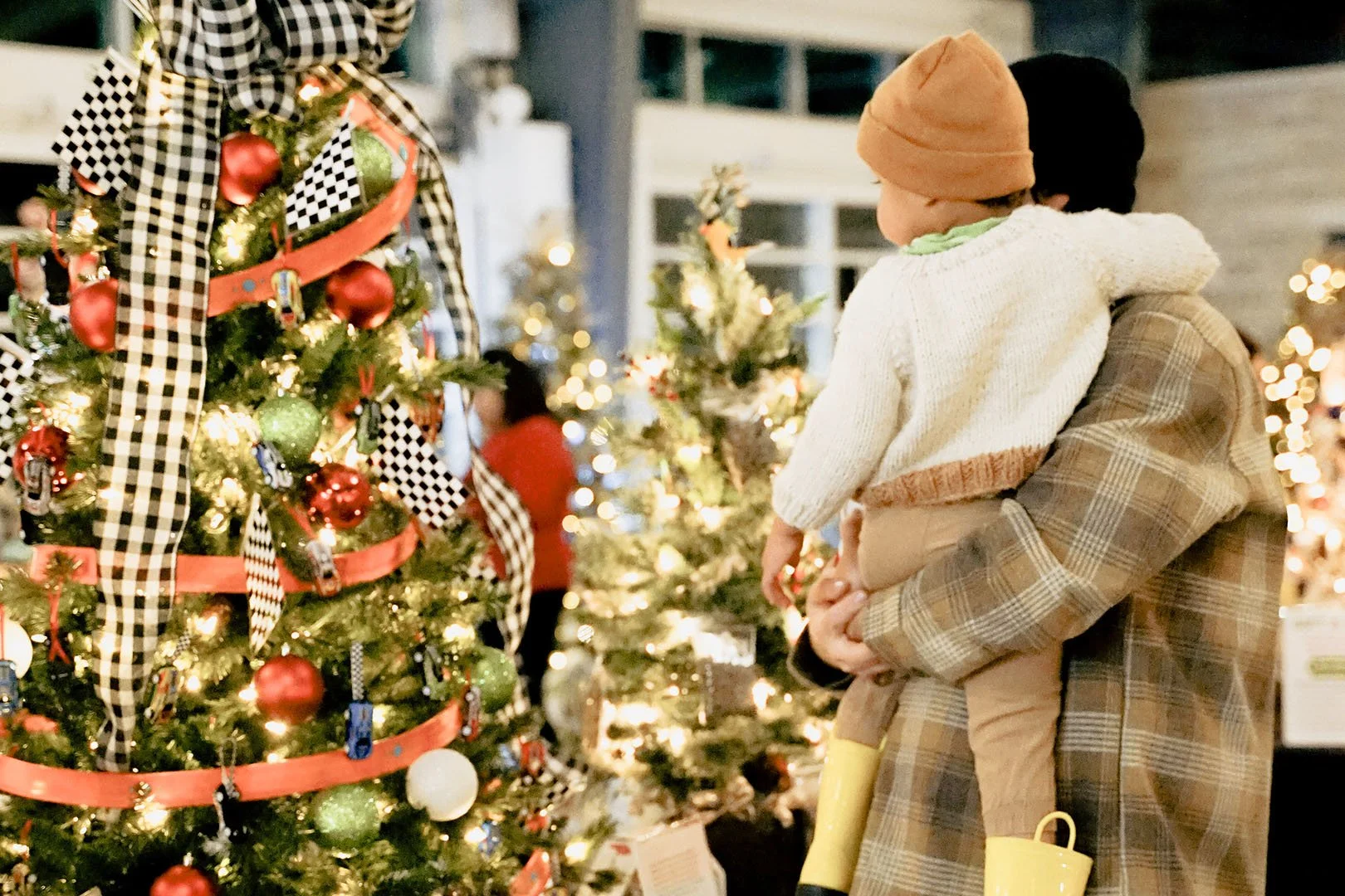 A person is holding a child while shopping for Christmas decorations in a store decorated with Christmas trees, lights, and ornaments.