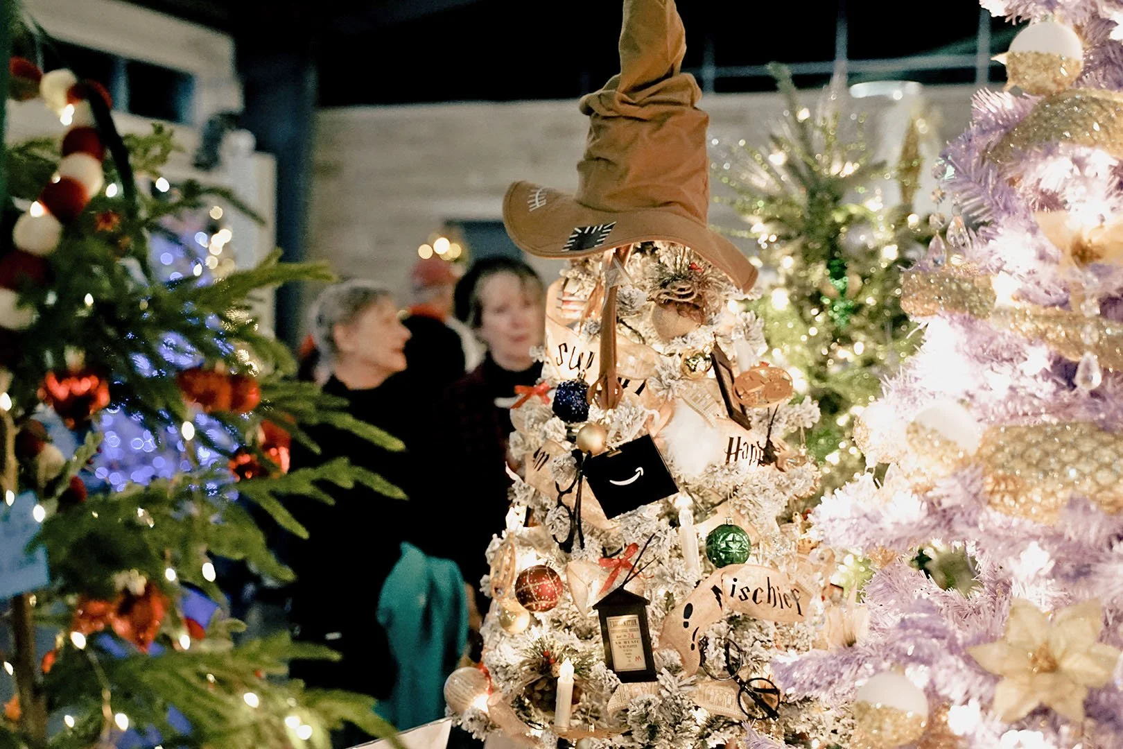 Decorated Christmas trees with ornaments, lights, and a witch hat on a white tree in the foreground. People are in the background at a holiday event.