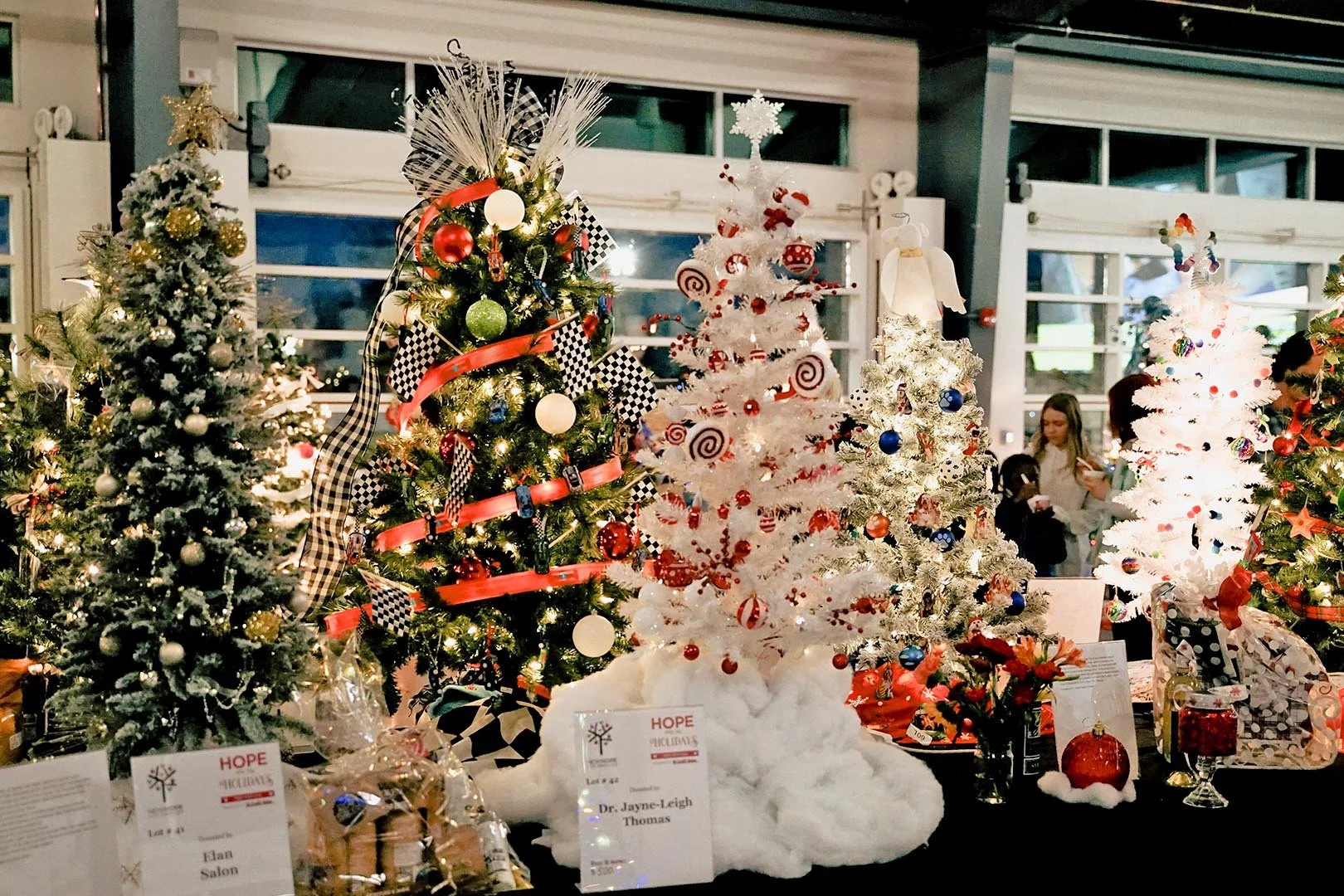 Various decorated Christmas trees on display, including a white tree with red and white ornaments, a white tree with multicolored decorations, and a snow-dusted tree with gold and silver ornaments. People are browsing in the background.
