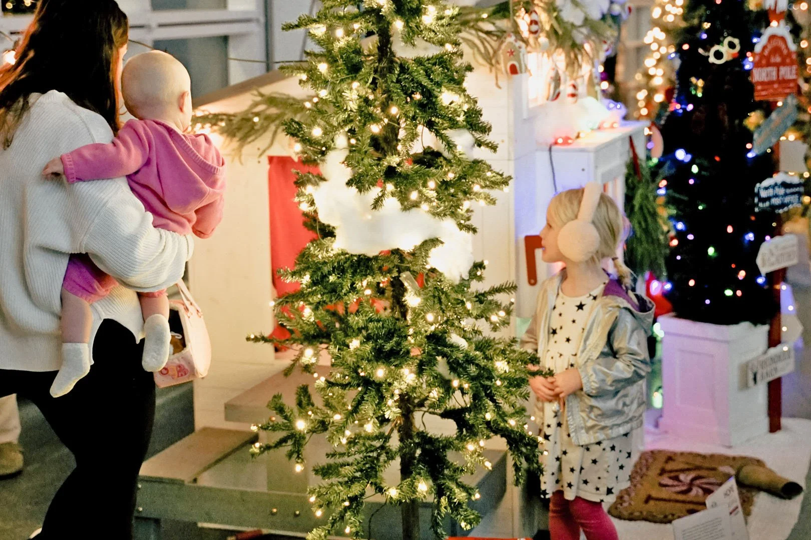 A woman holding a baby girl in front of a decorated Christmas tree, with a young girl wearing earmuffs looking at the tree in a festive store