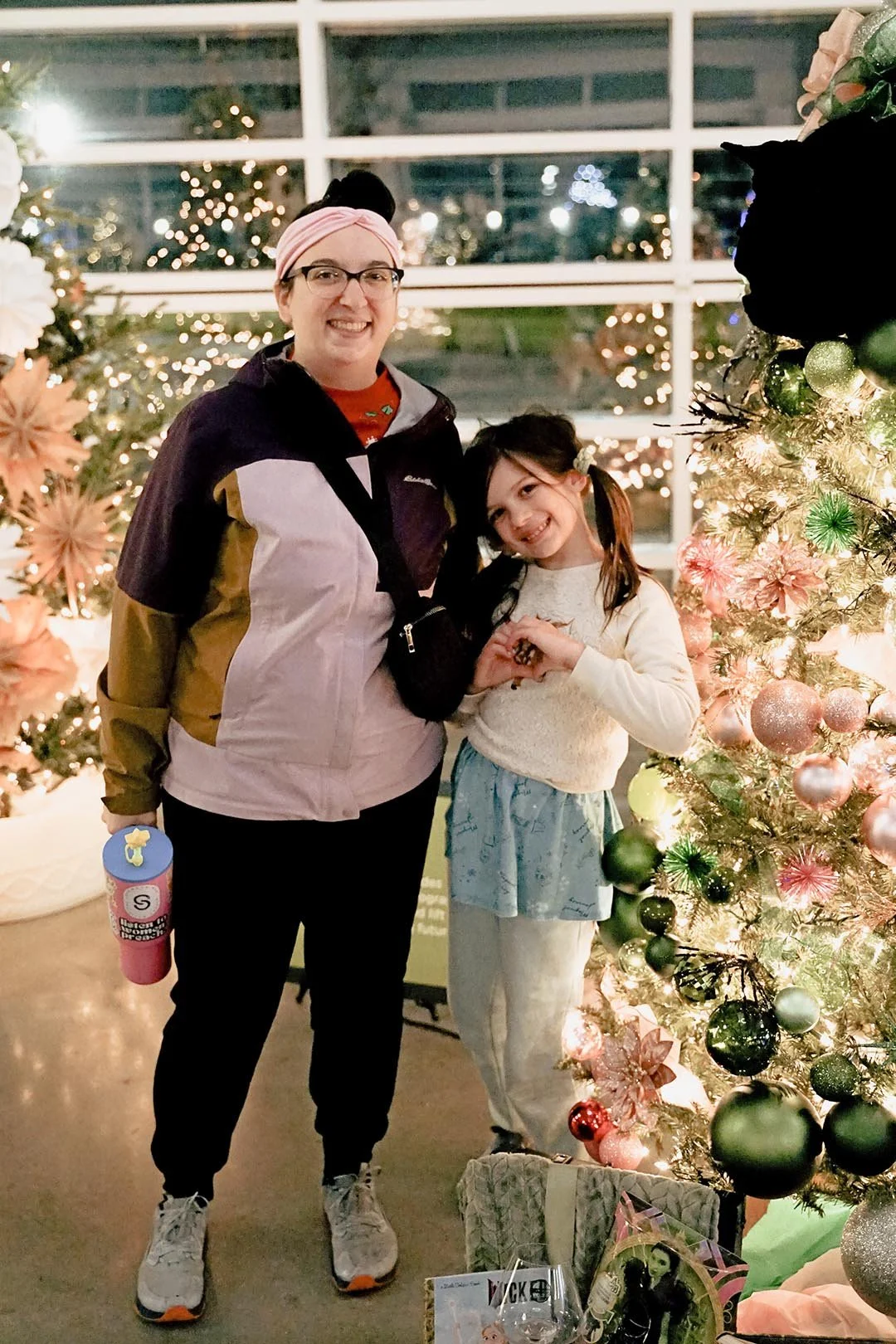Two females, one adult and one child, smiling and posing with a Christmas tree decorated with pink, green, and silver ornaments in a festive setting with other decorated trees and holiday lights in the background.