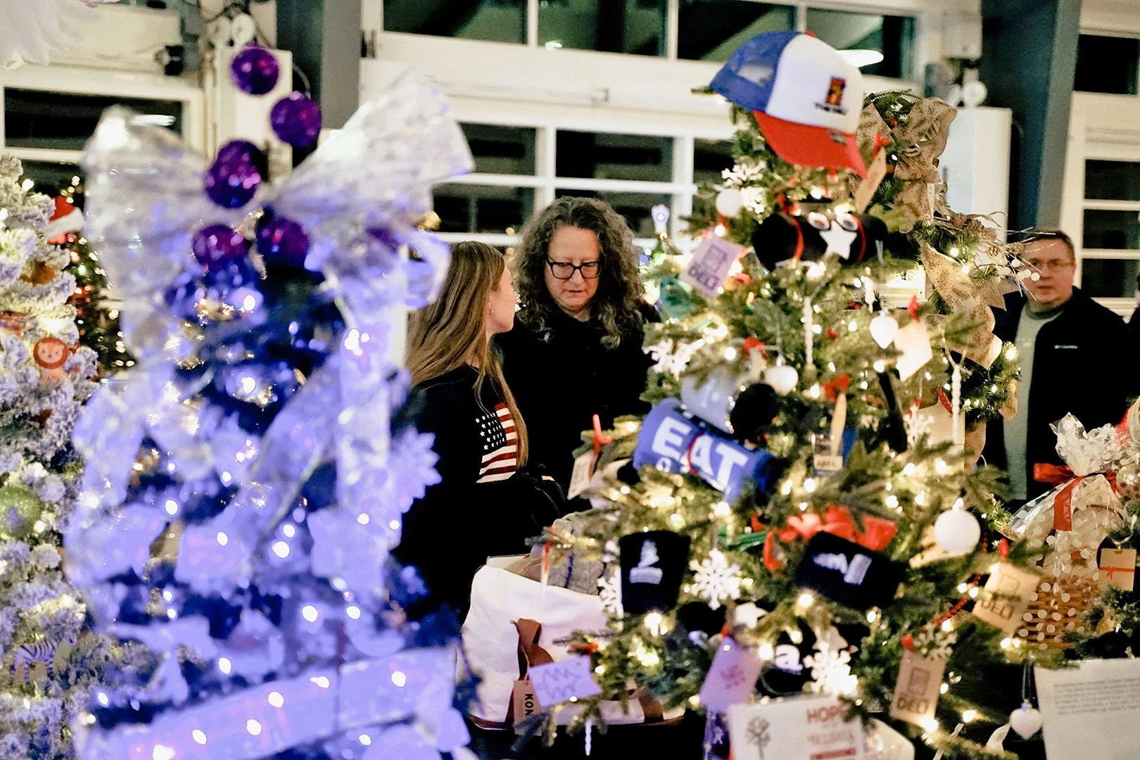 People shopping for Christmas ornaments at a decorated holiday display, with Christmas trees adorned with various ornaments and lights.