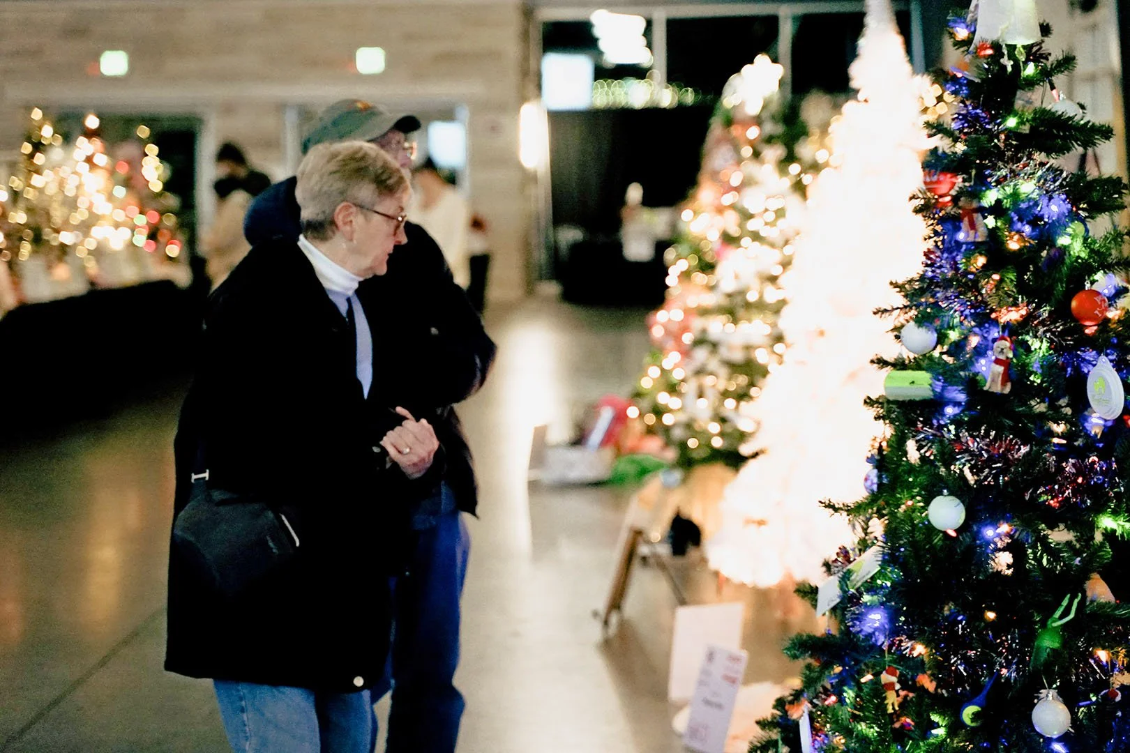 People viewing decorated Christmas trees in an indoor setting with soft lighting.