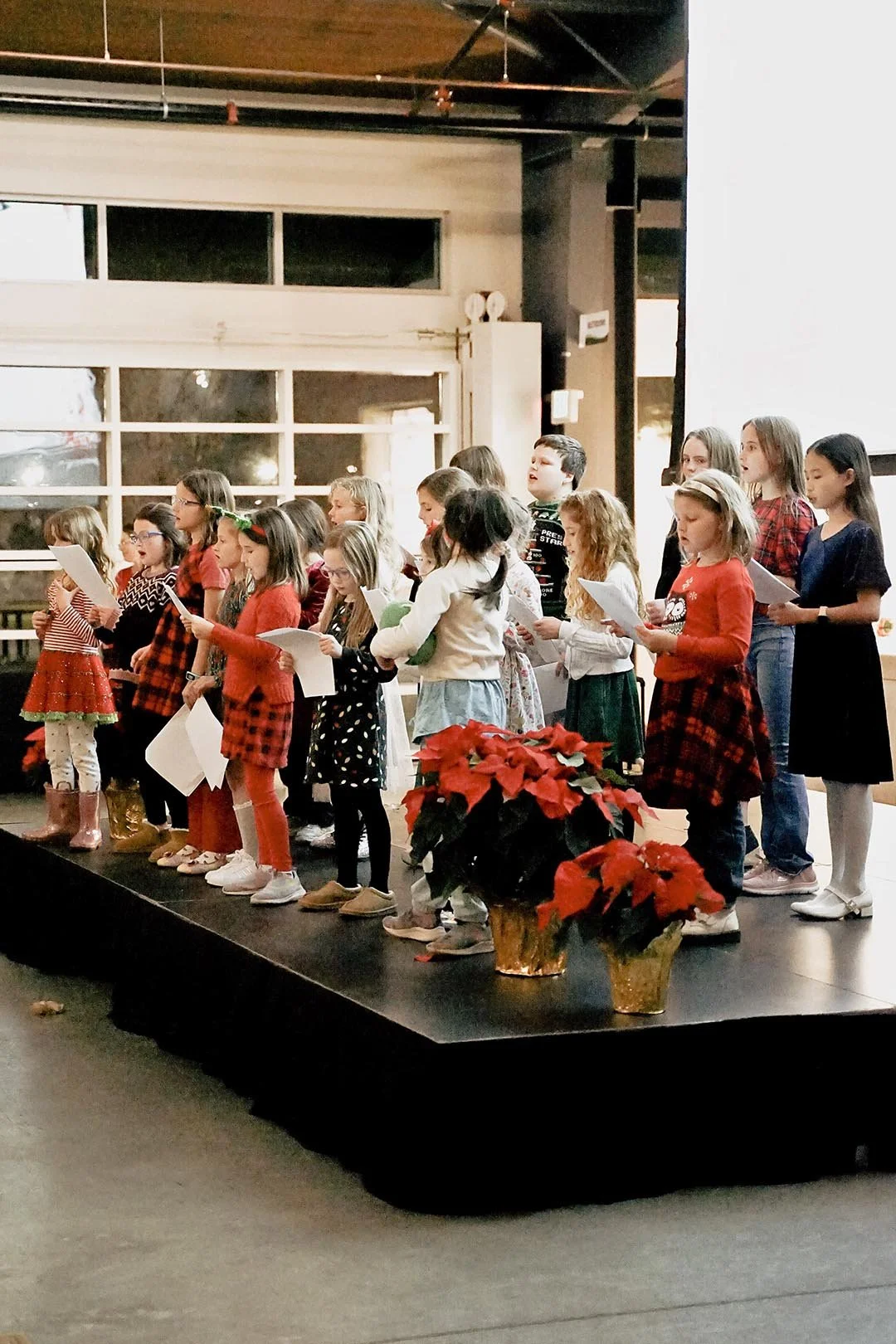 Children singing on a stage decorated with poinsettia plants, in a festive setting.