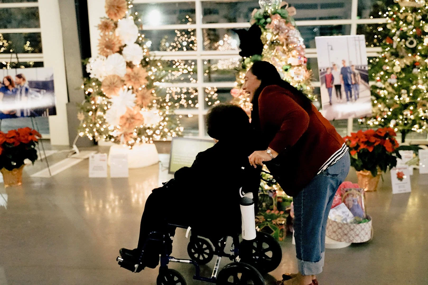 A woman leaning over a person in a wheelchair, both laughing, in front of decorated Christmas trees with lights, poinsettia plants, and holiday photos displayed.