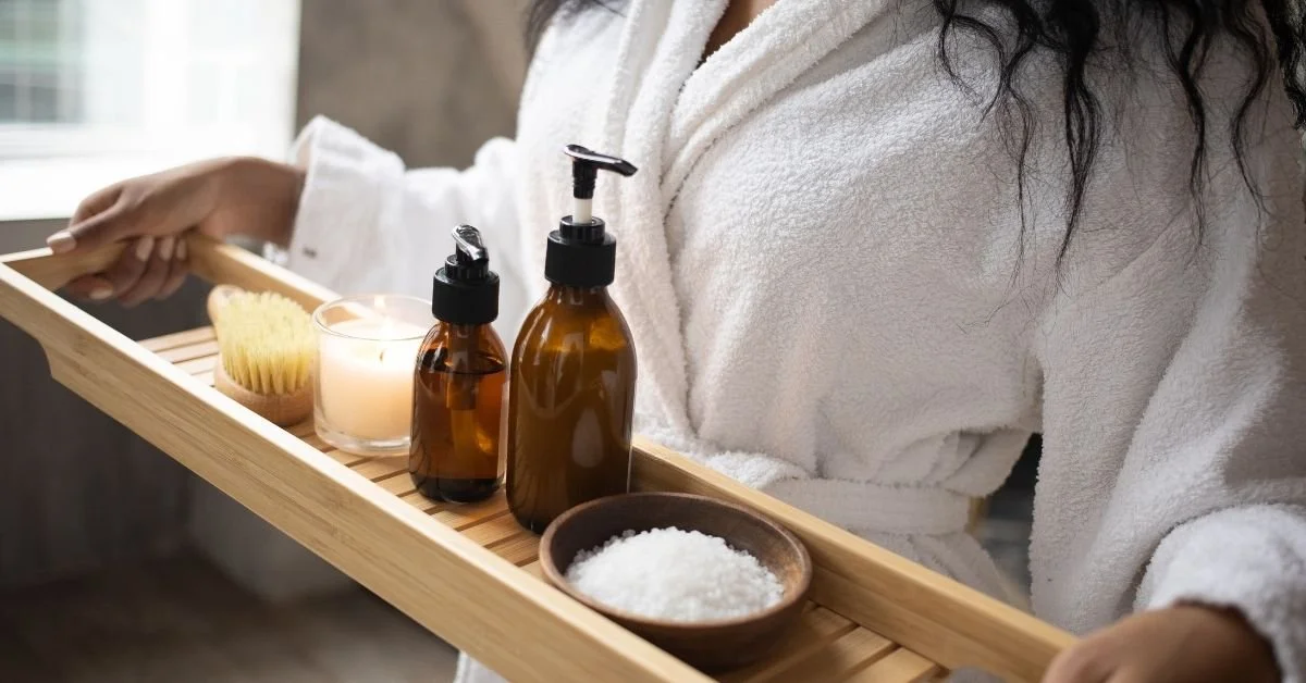 Person holding a wooden tray with bath essentials, including a brush, candle, amber bottles, salt, and lotion, in a cozy bathroom.