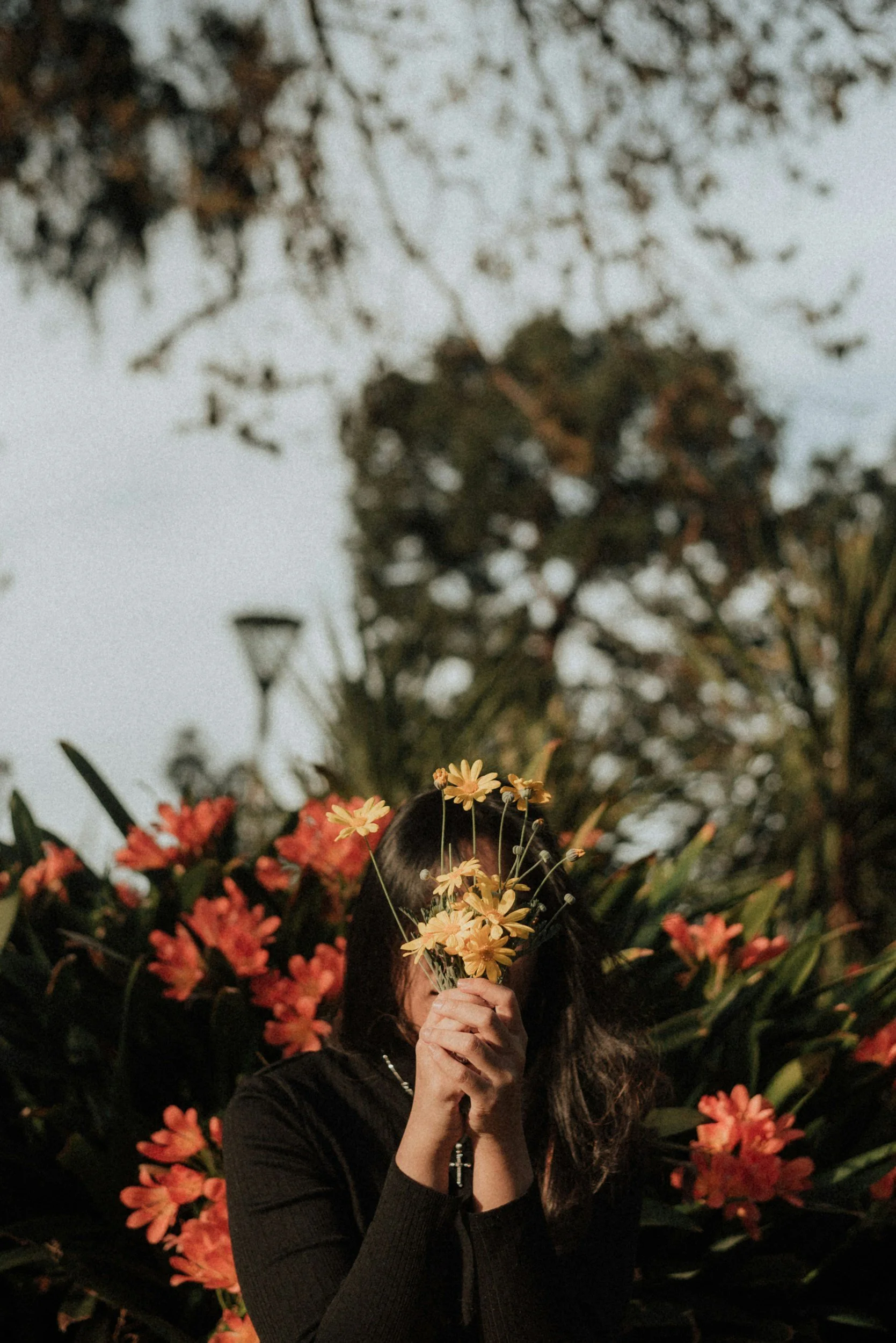 A woman with long dark hair holding a bunch of yellow flowers in front of her face, surrounded by red and orange flowers and greenery, with a background of trees and sky.