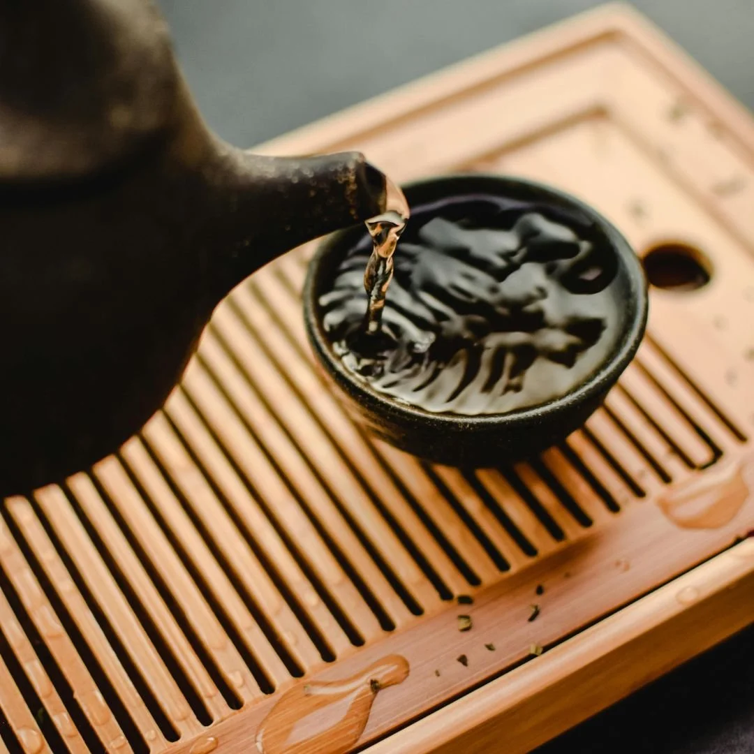 Close-up of black tea being poured from a teapot into a black cup on a wooden tray with a slatted design.