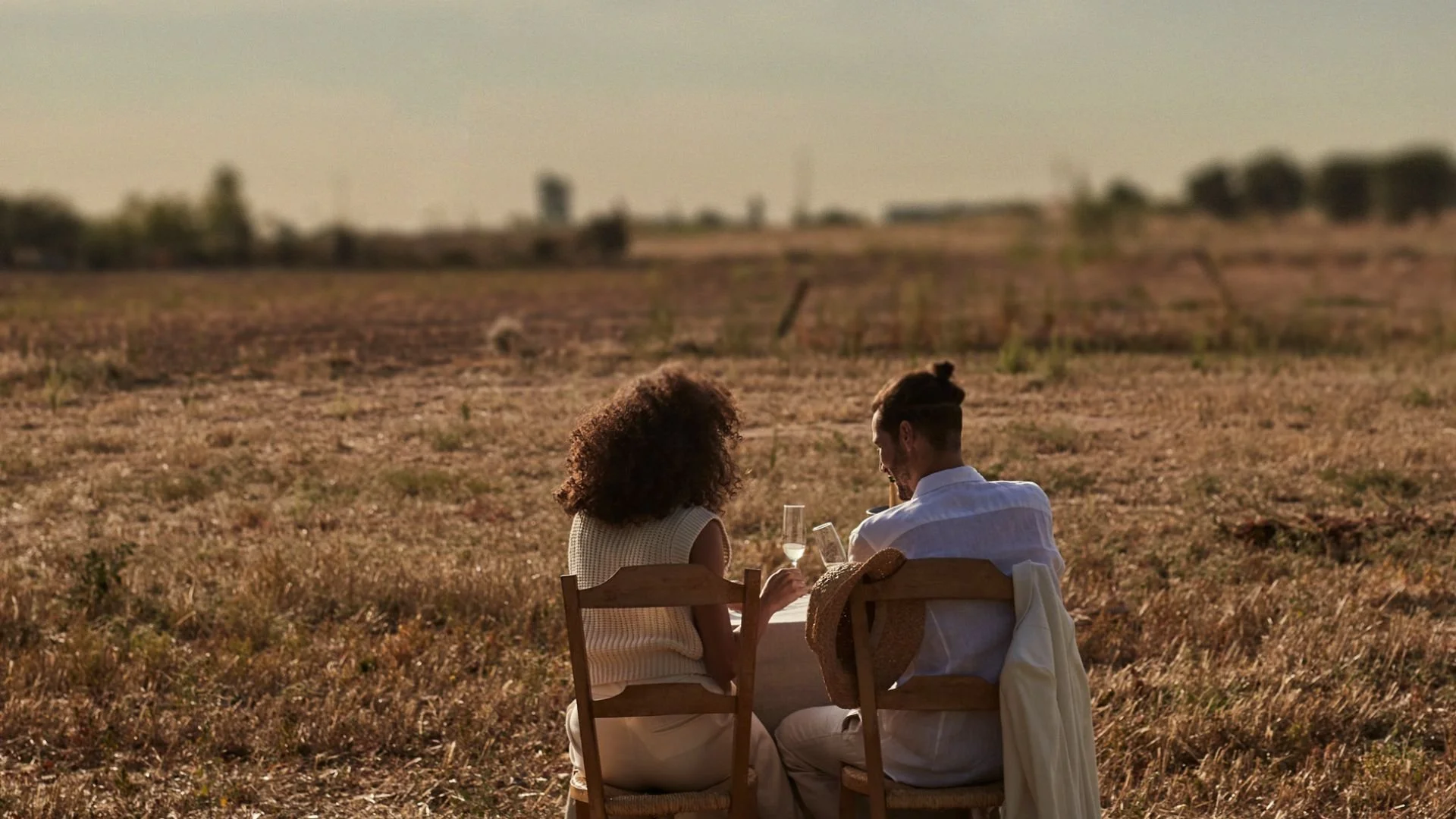 A couple sits at a table in a field, enjoying drinks and looking at their phones during sunset.
