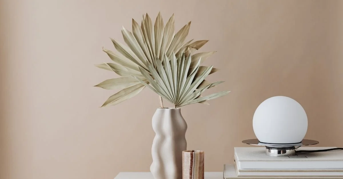 Decorative beige vase with a large dried palm leaf, white round lamp on a stack of books on a white surface against a beige wall.