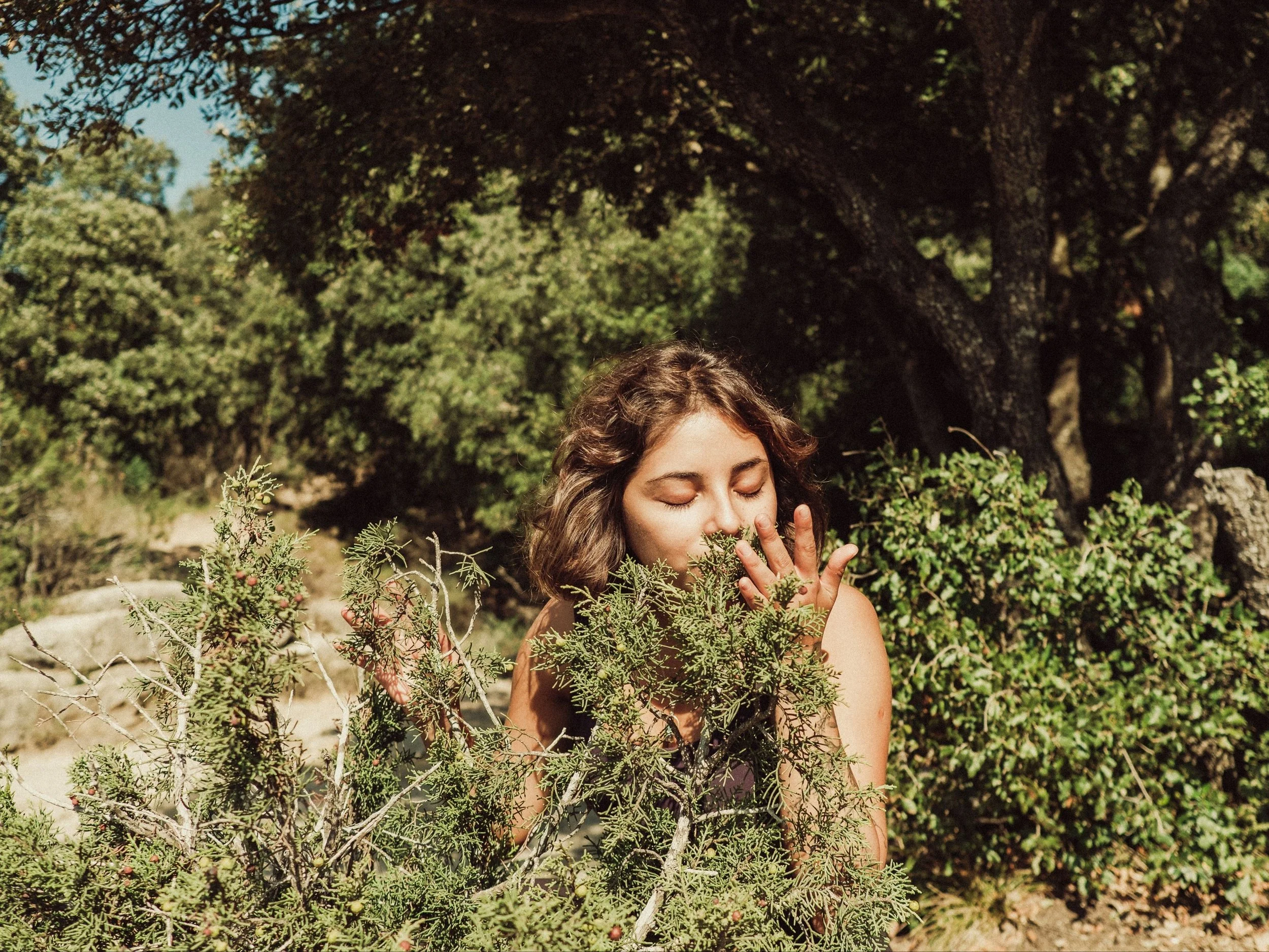 A young woman with curly hair is outdoors in a natural setting, with her eyes closed and nose near green foliage. She appears to be smelling or enjoying the surroundings. The background includes trees and shrubs, indicating a forest or park environment.