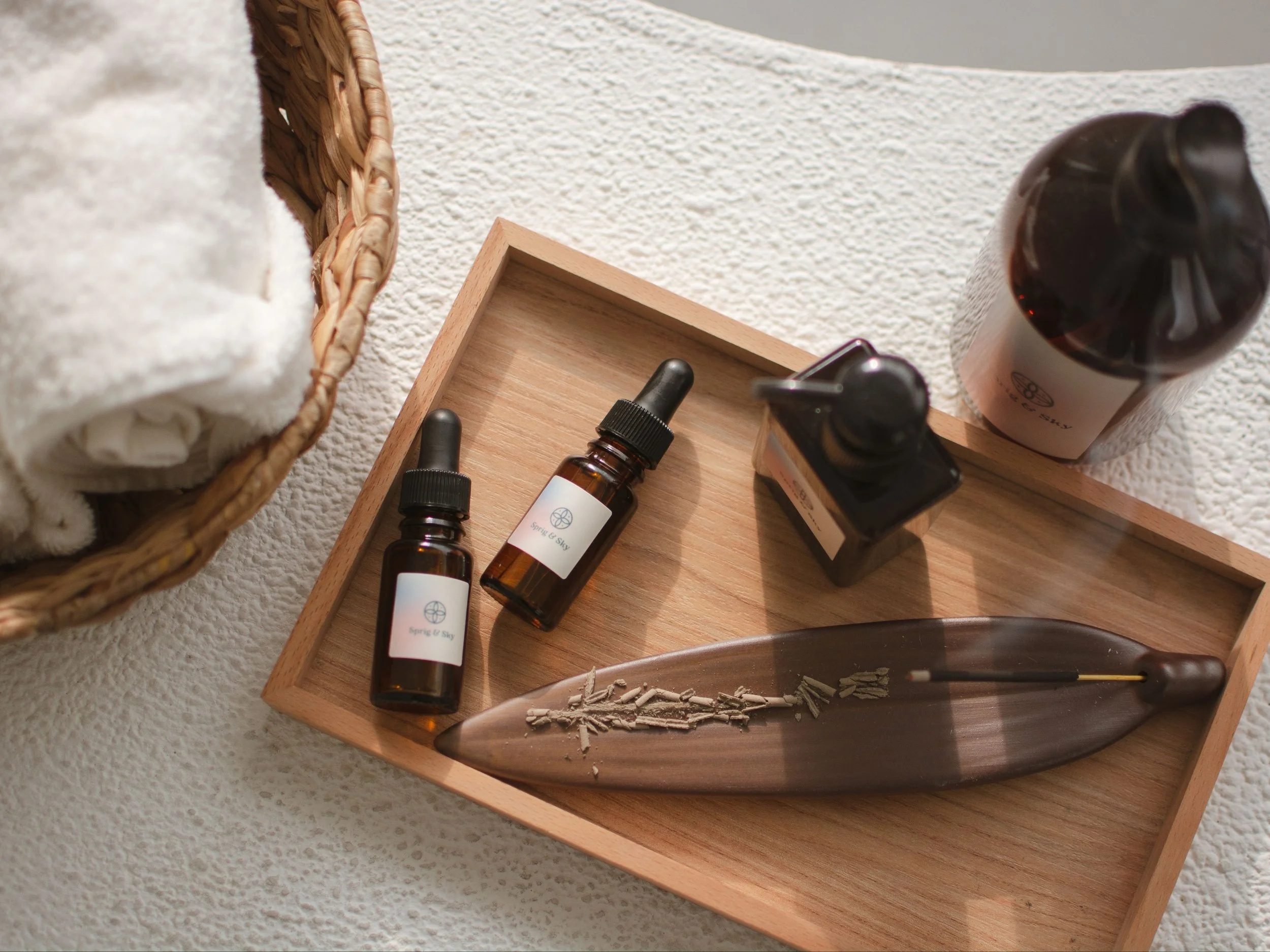 Wooden tray with two small bottles, one glass jar, a ceramic oil diffuser, and a wooden leaf-shaped object on textured surface. Basket with towel is partially visible.