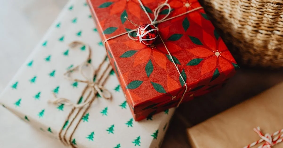 Two wrapped Christmas presents, one with red and green leaf pattern and the other with green trees pattern, decorated with twine and a small holiday ornament, placed on a surface near a wicker basket.