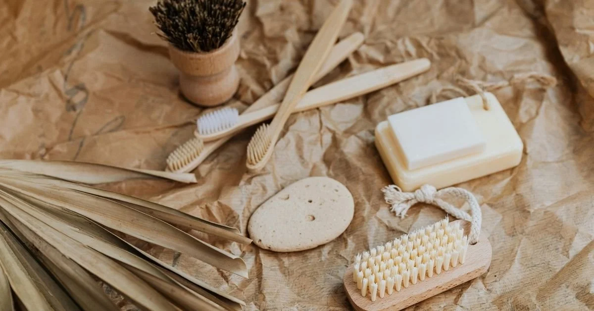 Natural skincare and hygiene items including soap, bamboo toothbrushes, a loofah, a soap dish, and dried leaves on crumpled paper background.