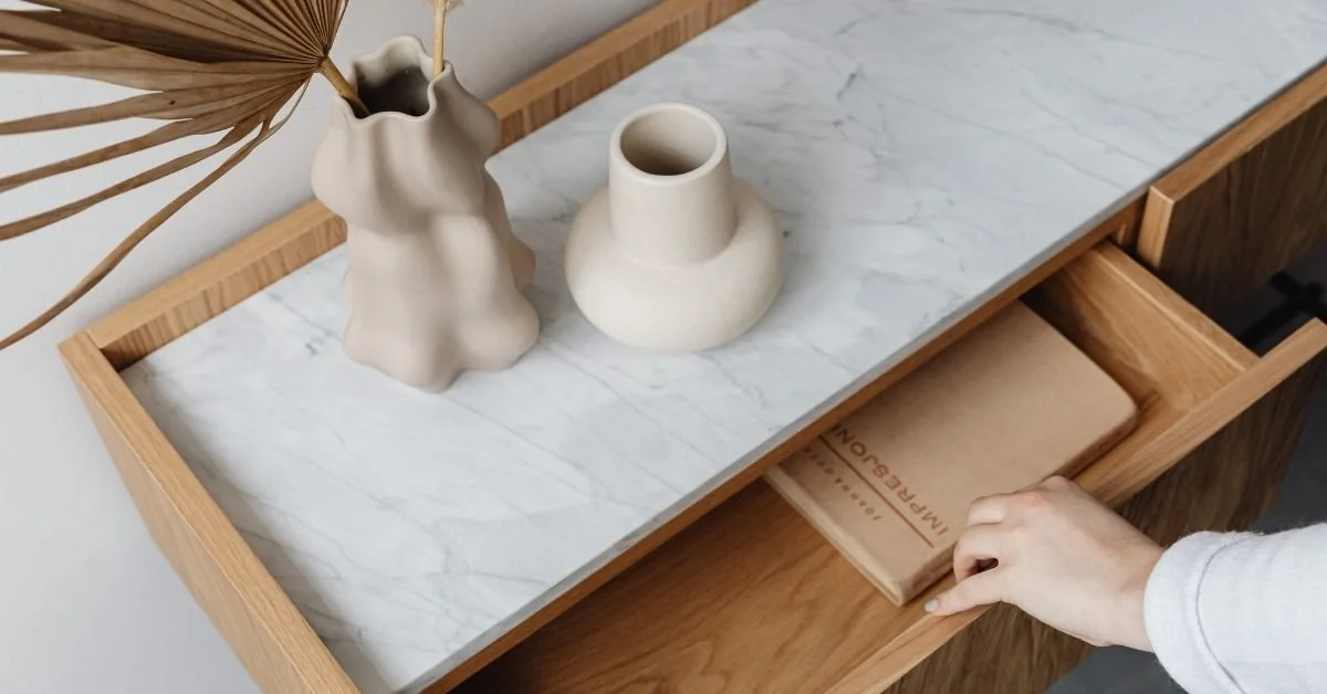 A wooden console table with a marble top, holding two ceramic vases, one with dried leaves, and a person pulling out a drawer with a notebook inside.