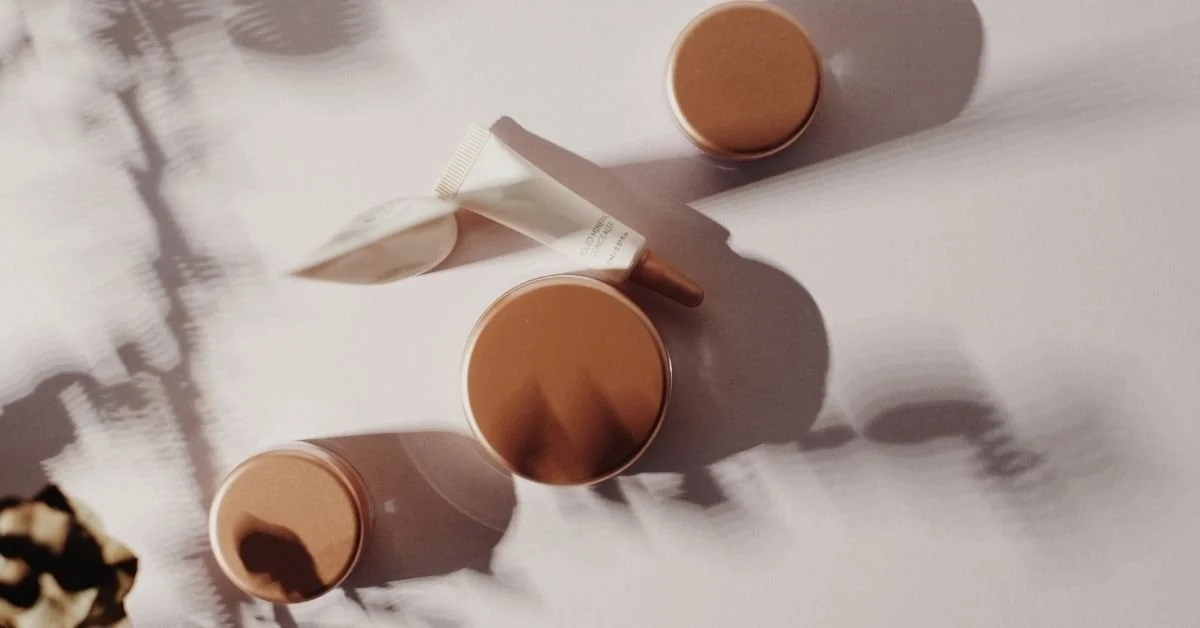 Close-up of three cups of coffee, a tube of cream, and a jar on a white surface, with shadows cast by natural light.