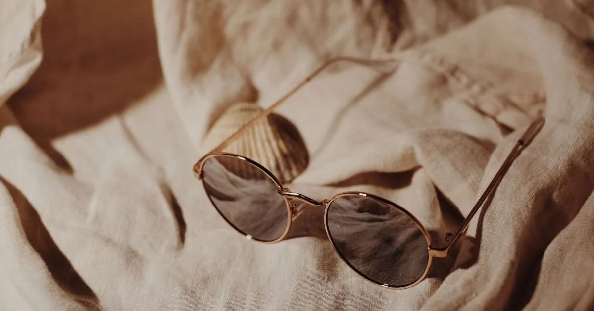 A pair of oval sunglasses with dark lenses and a thin metal frame resting on a beige fabric with a shell in the background.