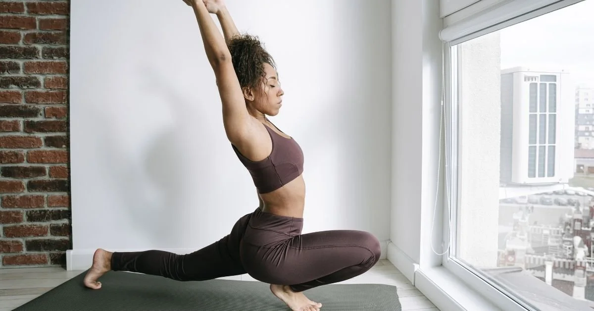 Woman practicing yoga in a lunging pose in front of a large window.