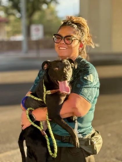 A woman with glasses and a ponytail smiles while holding a black dog with its tongue out, outdoors on a sunny day.