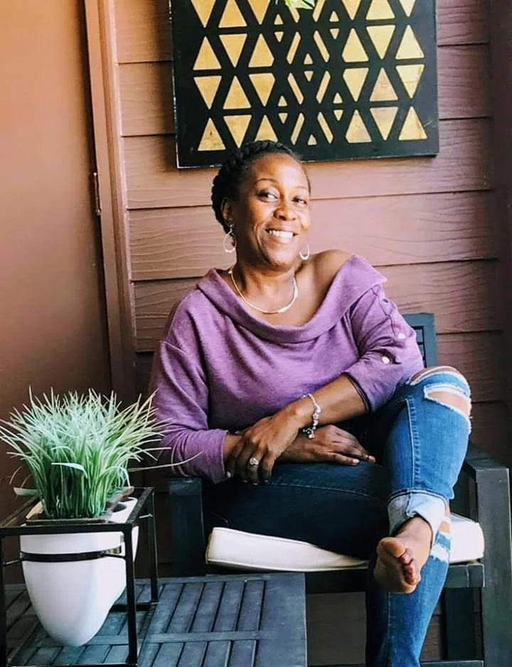 Woman sitting on a patio chair wearing a purple sweater and ripped jeans, next to a small table with a plant, in front of a wall with a geometric art piece.