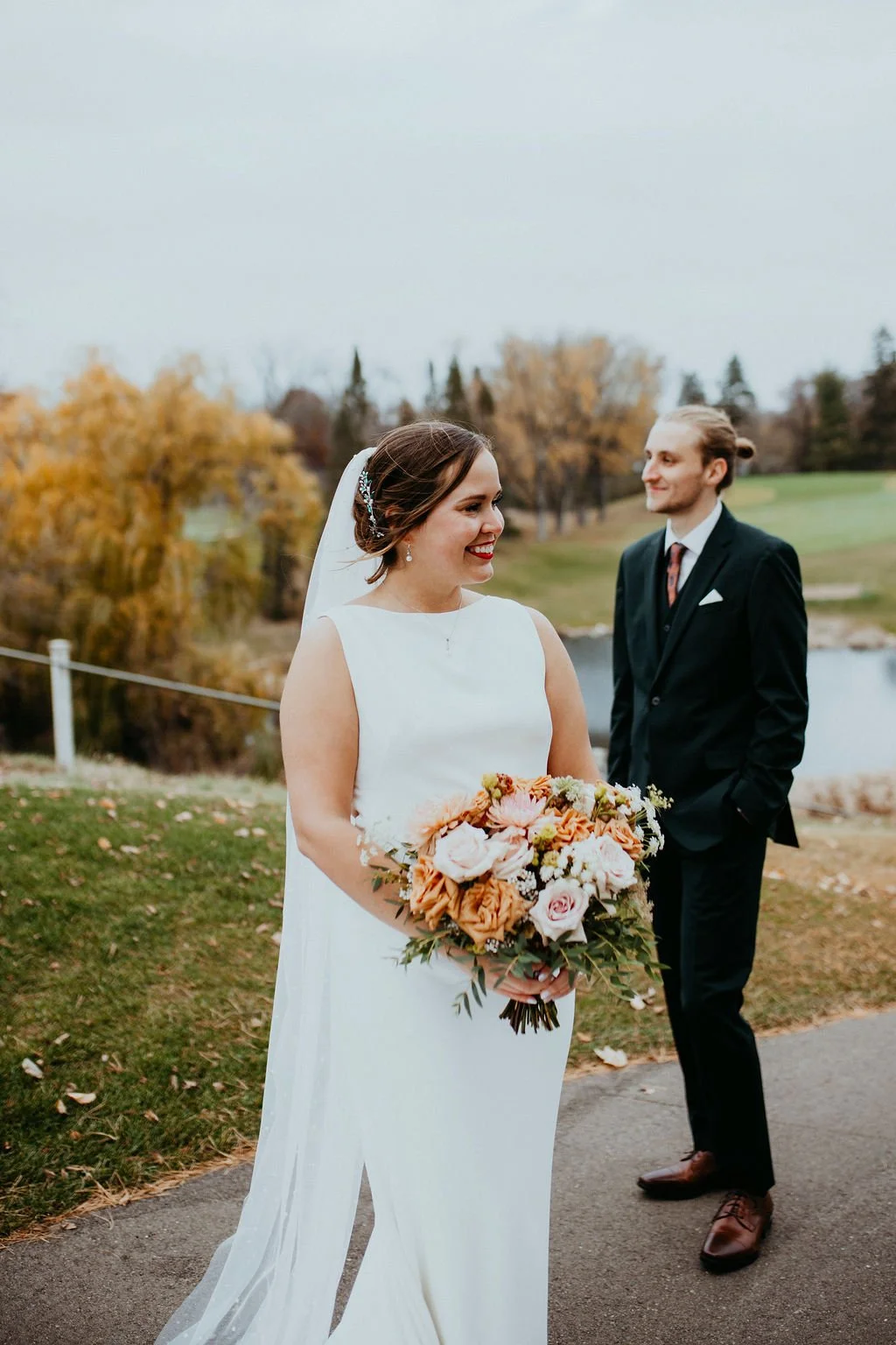 A bride in a white wedding dress holding a large bouquet of pink, peach, and white flowers, smiling while looking at a groom in a black suit with a white shirt and tie, standing outdoors on a cloudy day with autumn-colored trees and a lake in the background.