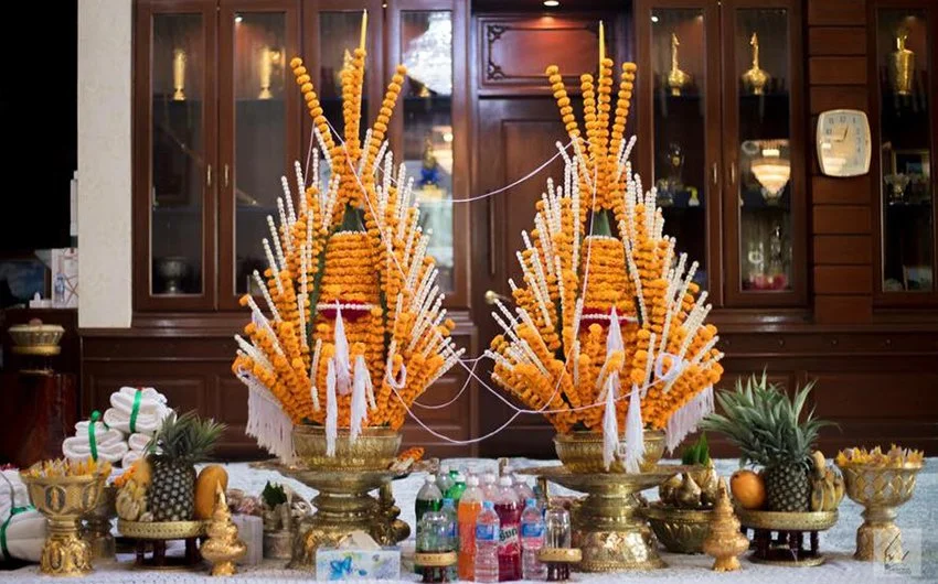 Two traditional Thai floral arrangements made of marigolds and white flowers on golden stands, surrounded by pineapples, oranges, and bottled drinks, set on a table in front of a wooden cabinet.