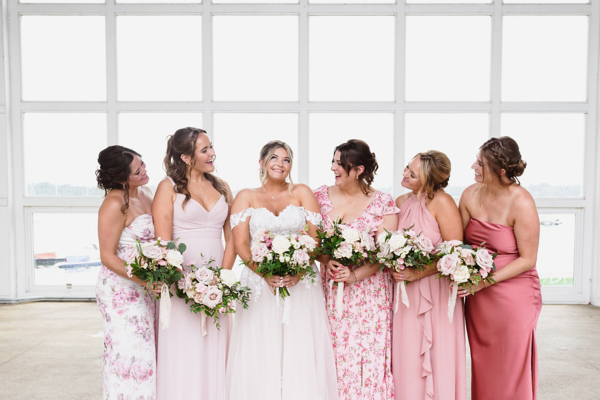 A bride and six bridesmaids standing together indoors in front of large windows, holding floral bouquets and smiling at each other.