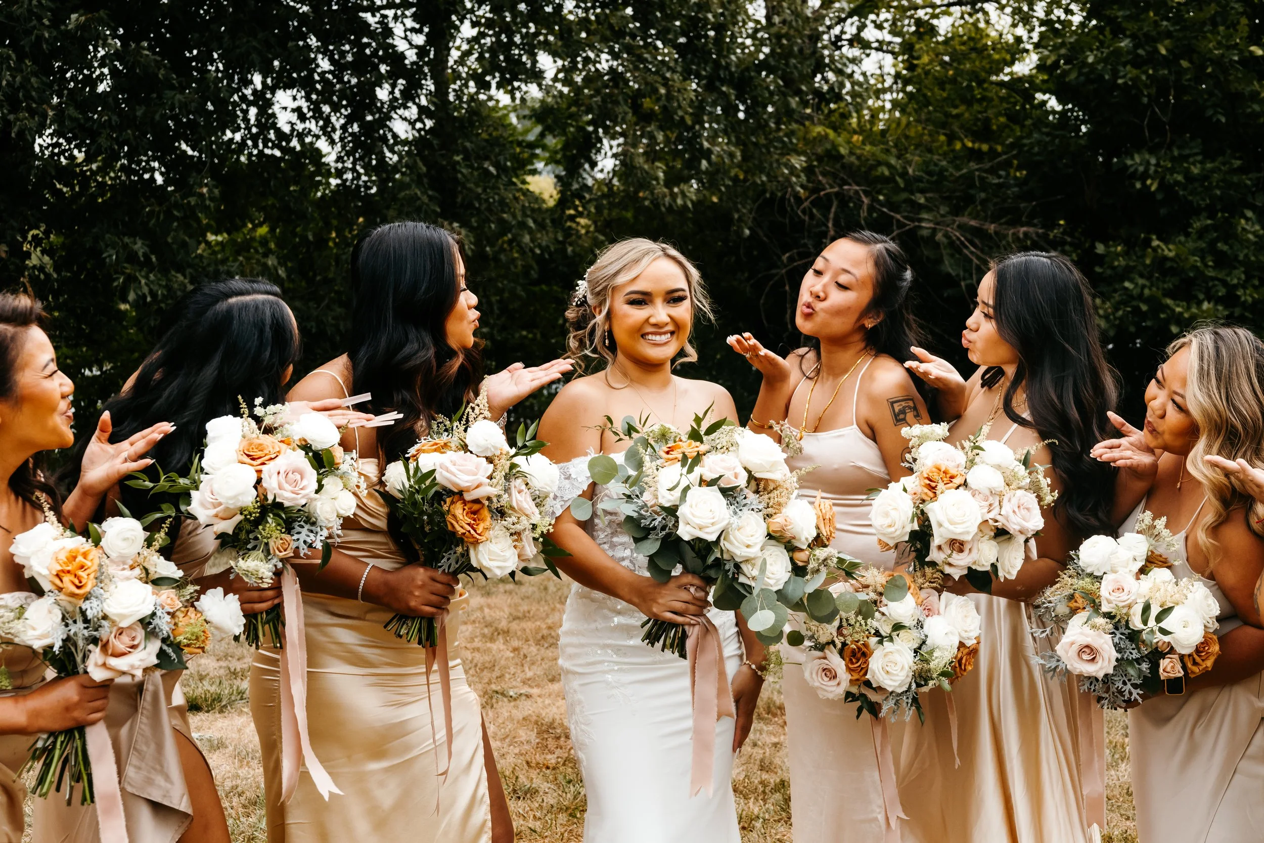 A group of women dressed in beige and white, holding bouquets of flowers, gathered outdoors for a wedding celebration. The bride in a white dress is smiling in the center, surrounded by friends.