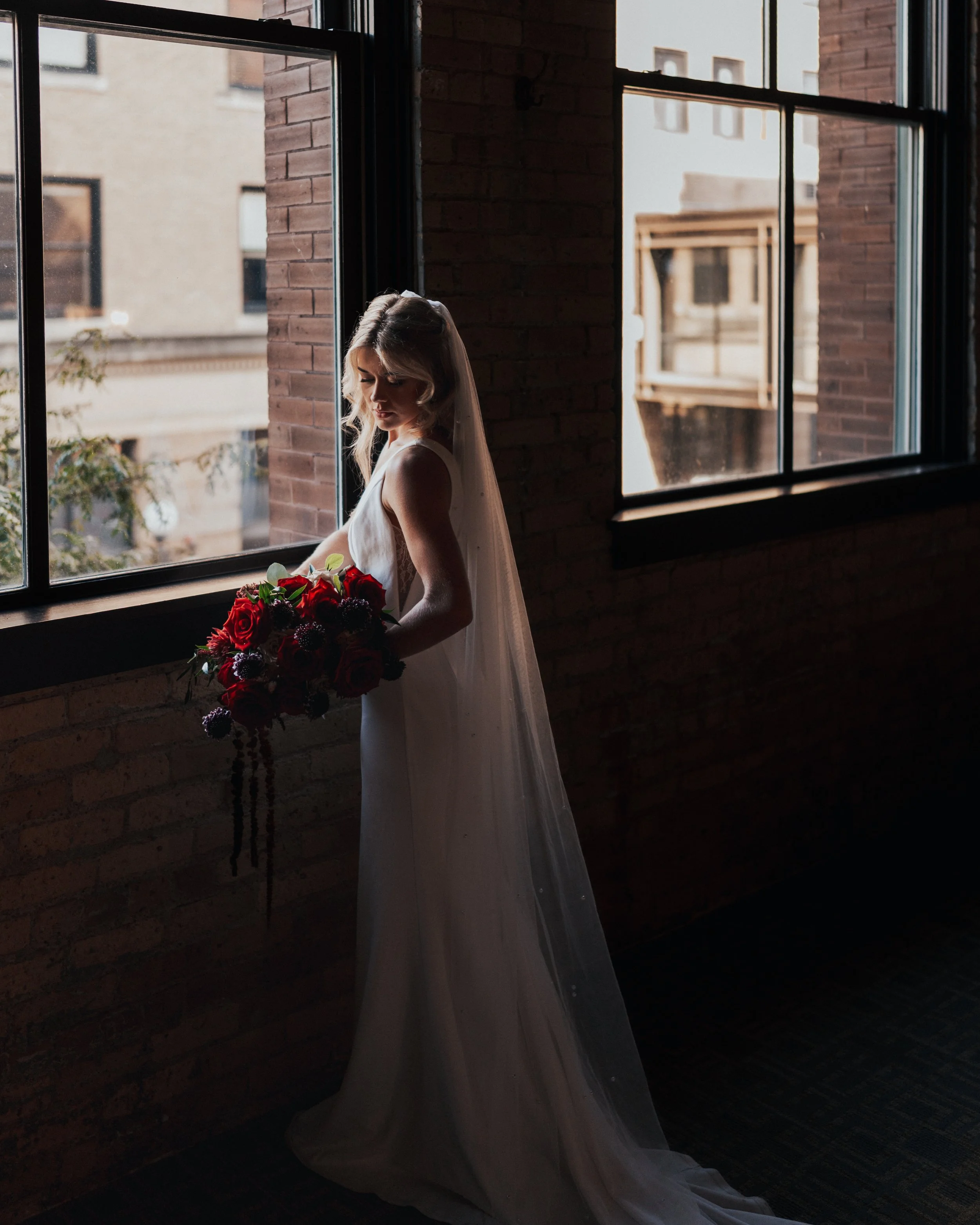 A woman in a white dress and veil holding a bouquet of red and purple flowers standing by a window with sunlight streaming through in an industrial-style brick interior.