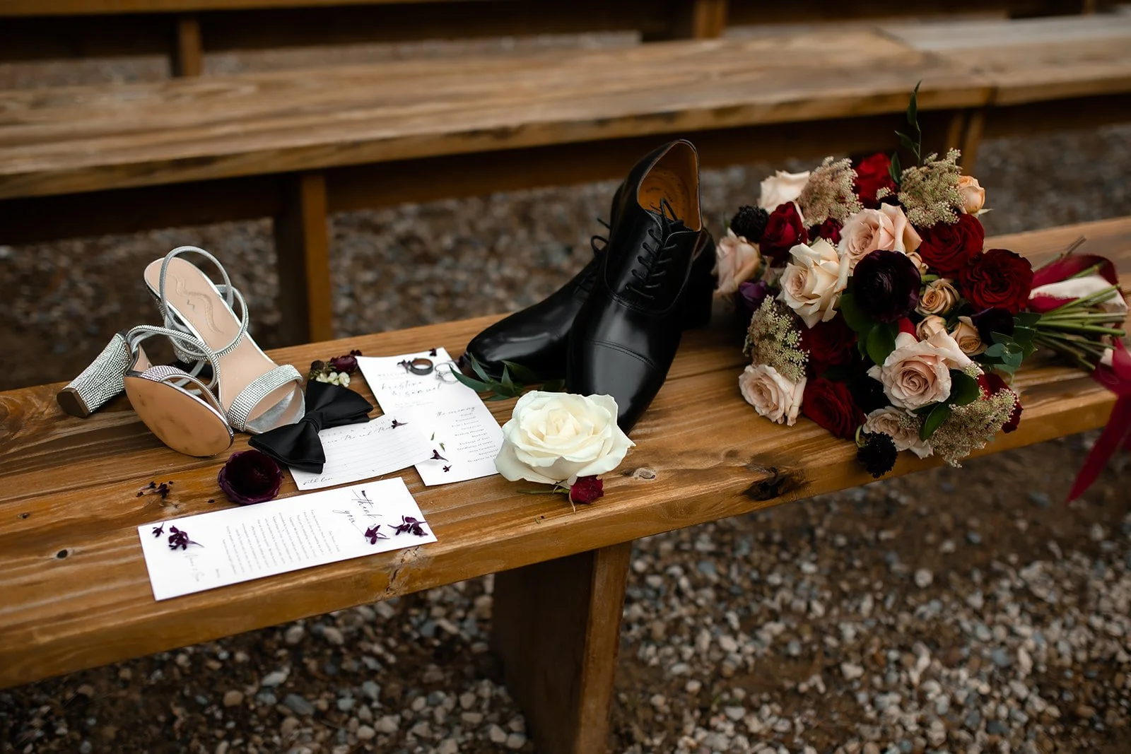 Wedding accessories and flowers on a wooden bench, including a pair of silver high-heeled sandals, black dress shoes, a bouquet of mixed roses and greenery, and wedding invitations.